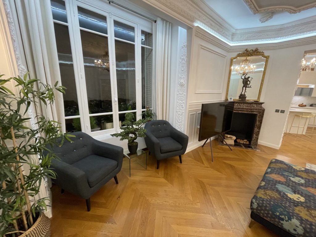 Salon haussmannien élégant avec parquet clair, moulures blanches et fauteuils gris.